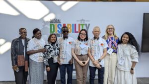 A diverse group of eight people, including youth delegates with disabilities and representatives from ADF, MasterCard Foundation, and Light for the World, pose in front of a banner for the Global Disability Summit 2025 in Berlin. Some wear white t-shirts with 'We Can Work' branding, while others wear professional or casual attire. A few participants have albinism, and some wear assistive devices like headsets or glasses. They are smiling and standing closely together.