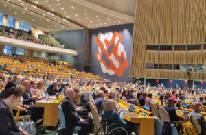 A wide view of the packed UN General Assembly Hall during COSP18, with delegates from around the world seated at rows of desks. The iconic orange and white mural is visible in the background, and several attendees, including wheelchair users, are engaged in the proceedings.